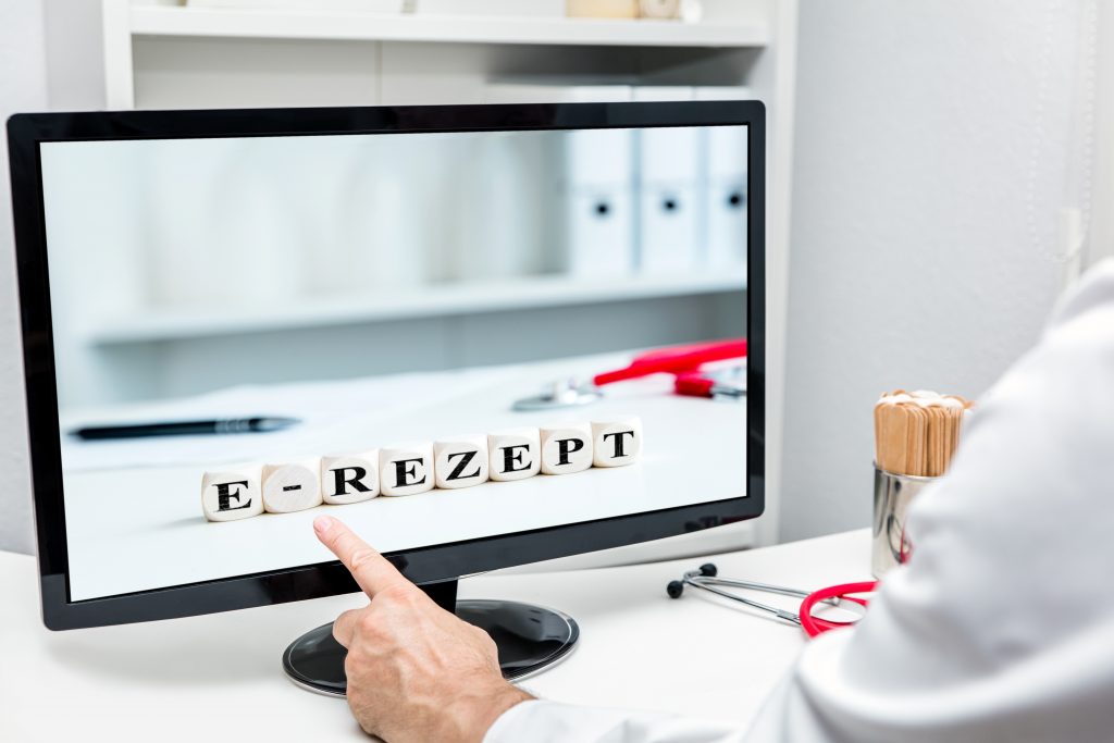 Auf einem PC-Monitor sind Holzwürfel mit dem Aufdruck E-Rezept zu sehen. Davor ein Arm mit weißem Kittel, ein Zeigefinger zeigt auf den Monitor.