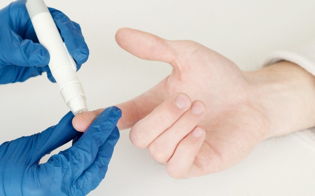 Hände in blauen Handschuhen stechen mit einem Insulinpen in den rechten Zeigefinger einer Hand, um das Blut eines Diabetikers zu kontrollieren.